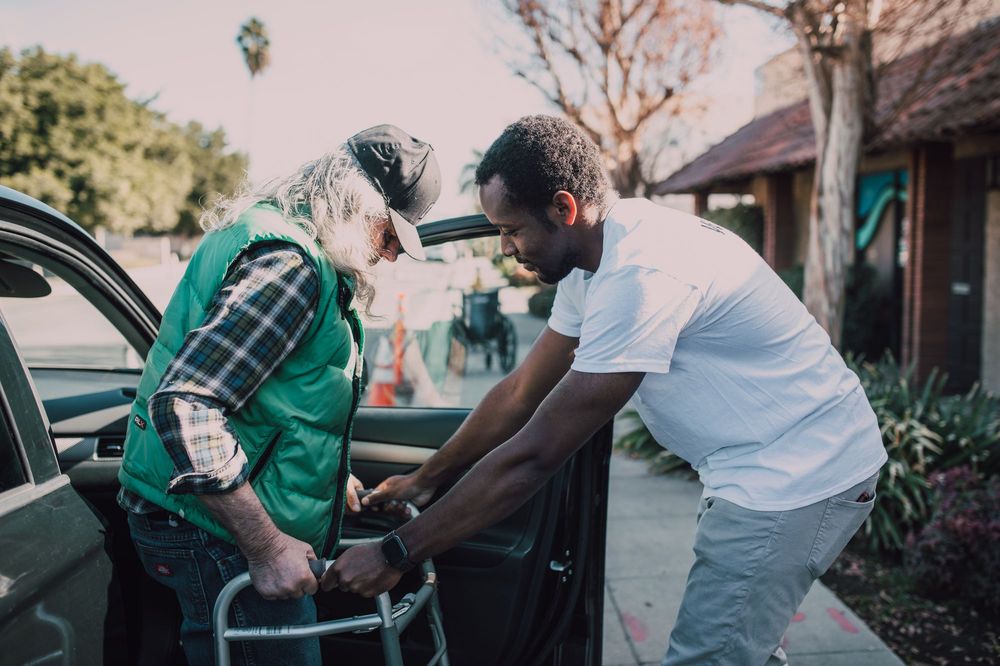 A young man helps an older man with a walker get out of a car.