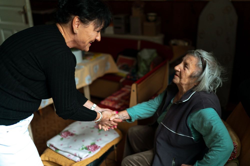 A caregiver warmly takes the hand of an older woman sitting in her living room.