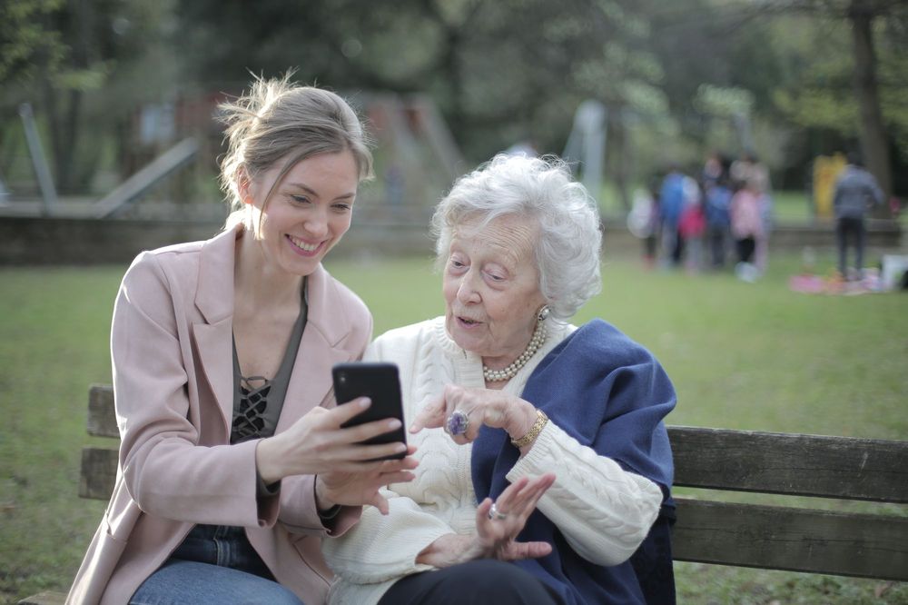 A young woman shows her smiling grandmother something on a phone while they sit on a park bench.
